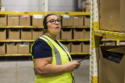 Mature female worker with digital tablet looking away while talking on headset at distribution warehouse