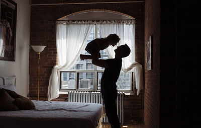Silhouette father picking up daughter while standing by bed at home