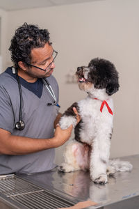 Hispanic man in uniform with stethoscope and glasses touching injured paw of obedient labradoodle dog with black and white fur during work in veterinary clinic