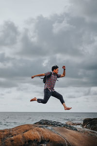 Man jumping above rock at beach against cloudy sky