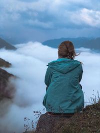 Rear view of woman sitting on cliff against cloudscape