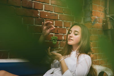 Young woman holding grapes while sitting on sofa against brick wall