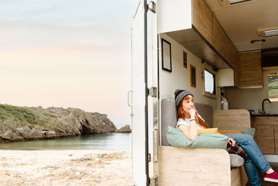 Side view of thoughtful traveling female hipster sitting on couch in truck and enjoying vacation near lake