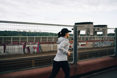 Young sportswoman jogging by fence on bridge in city