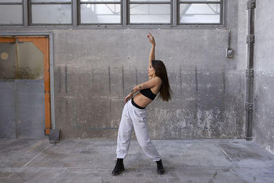 Female dancer dancing with arm raised against gray wall in abandoned factory