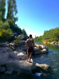 Tilt-shift image of shirtless man standing on rock at riverbank against blue sky