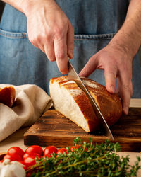 Faceless man cutting fresh home baked crusty bread with large knife on wooden board on kitchen 