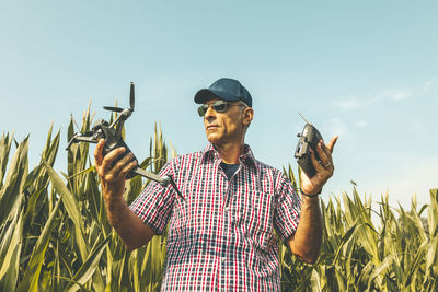 Low angle view of mature man wearing sunglasses holding drone at field against sky