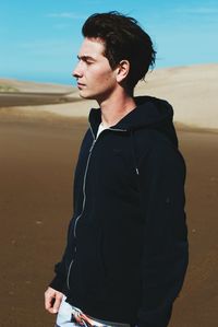 Side view of thoughtful young man looking away while standing on sand against blue sky