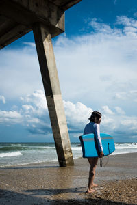 Side view of man with surfboard standing at beach against cloudy sky during sunny day