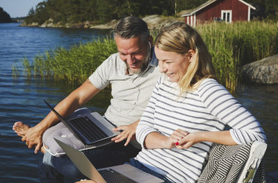 Smiling couple using laptop while sitting on bench against lake