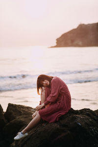 Portrait of woman sitting on rock at beach against sky