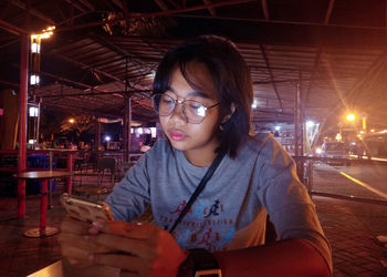 Young woman using phone while sitting at outdoor restaurant during night