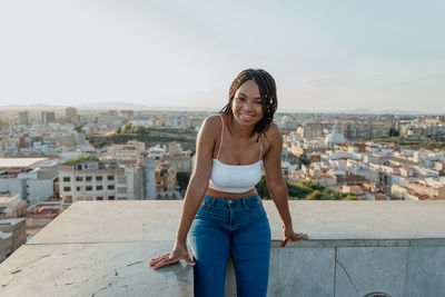 Content young african american female in jeans and crop top looking at camera on fence in city