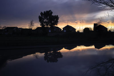 Reflection of silhouette tree and buildings by lake against sky during sunset