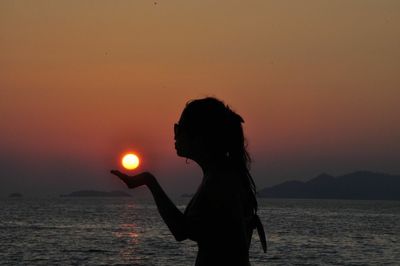 Optical illusion of silhouette woman holding sun at beach during sunset