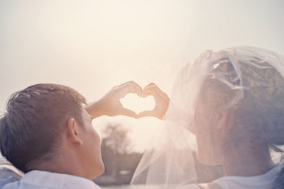 Rear view of newlywed couple forming heart shape against sky during sunny day