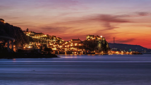 Illuminated buildings by sea against sky at sunset