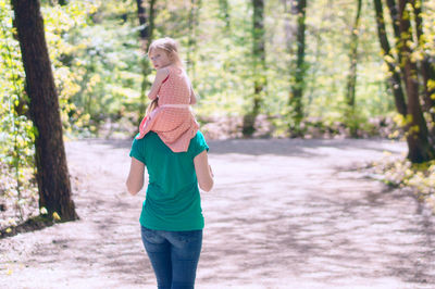 Rear view of woman carrying daughter on shoulder while walking in forest