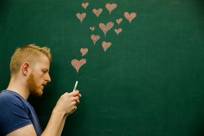 Young man using mobile phone with heart shapes on blackboard