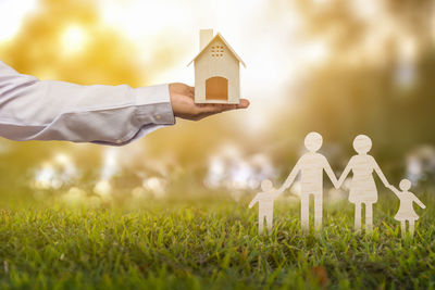 Cropped hand of man holding model home with family figurine on grassy field