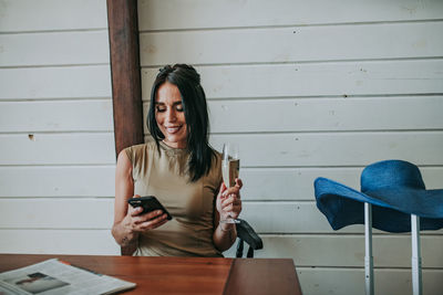 Woman using smart phone holding champagne flute sitting at home