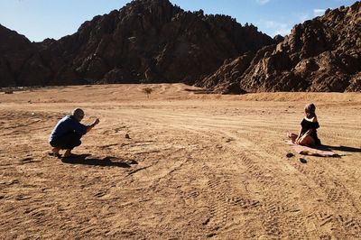 Side view of man photographing woman sitting on sand