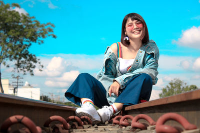 Low angle portrait of smiling young woman wearing sunglasses sitting on railroad track