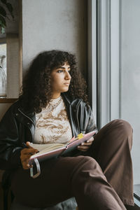 Thoughtful young woman sitting with book looking out of window at university