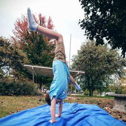 Boy practicing handstand on blue fabric at grassy field against sky