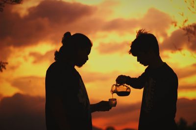 Man pouring drink for friend against sky during sunset