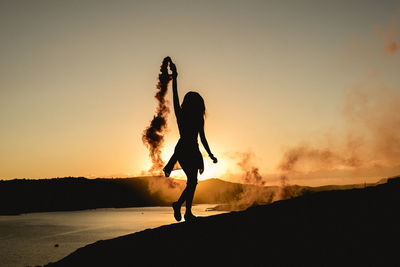 Silhouette woman holding distress flare while walking against sea during sunset
