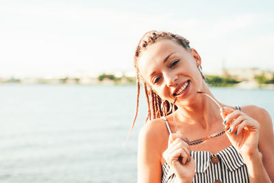 Close-up portrait of smiling young woman holding sunglasses against lake