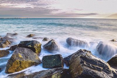 Scenic view of waves crashing on rocks against cloudy sky