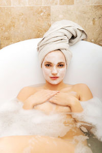 Portrait of naked young woman with facial mask lying in bathtub at home