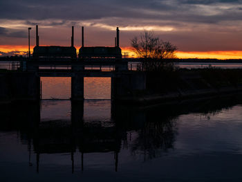 Silhouette factory by lake against sky during sunset