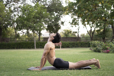 Side view of shirtless man sitting on grass against trees