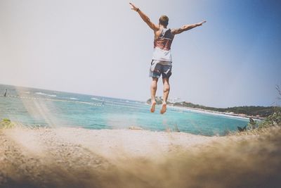 Rear view of man jumping with arms outstretched at beach against sky