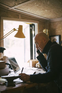 Businessman talking over mobile phone while using laptop in workshop