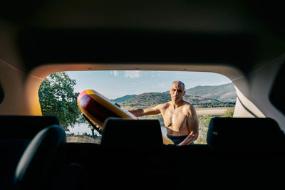 Active middle aged shirtless man taking paddle board from automobile parked on lake shore in hilly nature while preparing for water sport practice in summer day