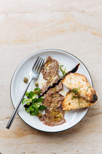 Top view of tasty fried pieces of meat on white plate with roasted bread decorated with sauce greenery and metal fork in restaurant