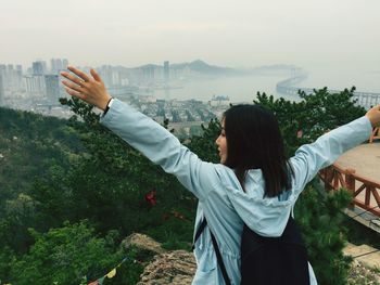 Rear view of young woman with arms outstretched standing on mountain against sky in city