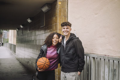 Portrait of smiling girl holding basketball while standing with male friend near underpass