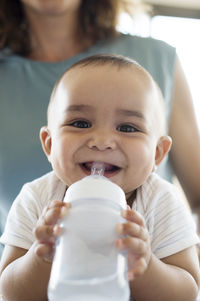 Portrait of smiling baby boy holding bottle while sitting with mother