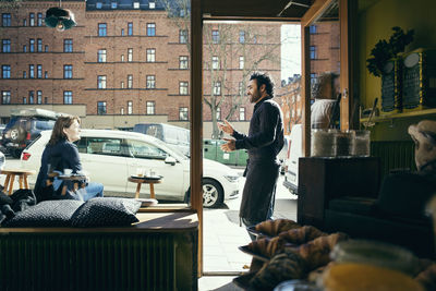 Smiling waiter talking with female customer on sidewalk seen through glass at cafe