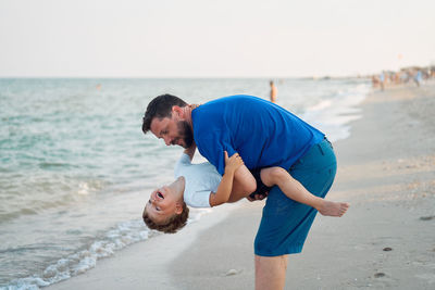 Young man carrying cheerful son at beach against clear sky