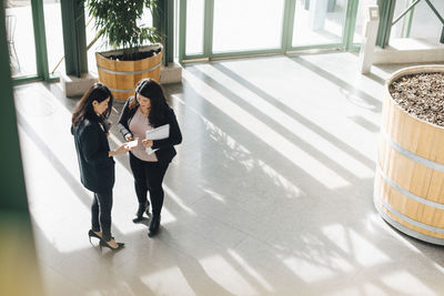 Female coworkers discussing over smart phones while standing at office corridor