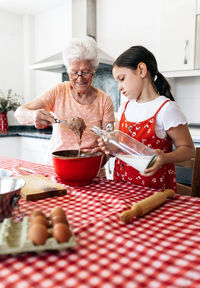 Grandmother in eyewear with whisk near granddaughter with bottle of milk cooking dough at table in house