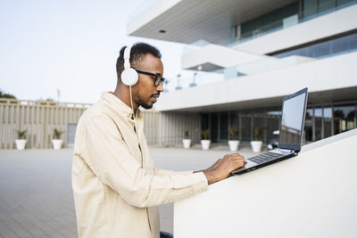 Freelancer wearing headphones using laptop outside building