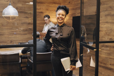 Portrait of cheerful businesswoman standing with laptop and diary at board room doorway in office